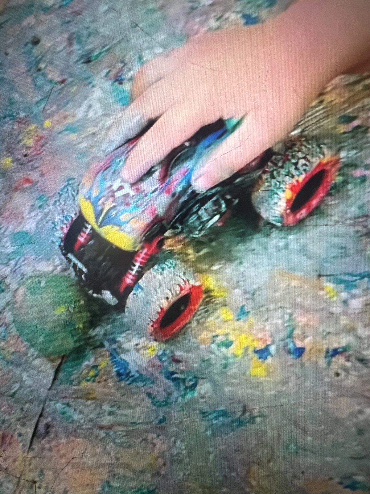 A child's hand pushing a paint-covered toy car across a colourful painted surface